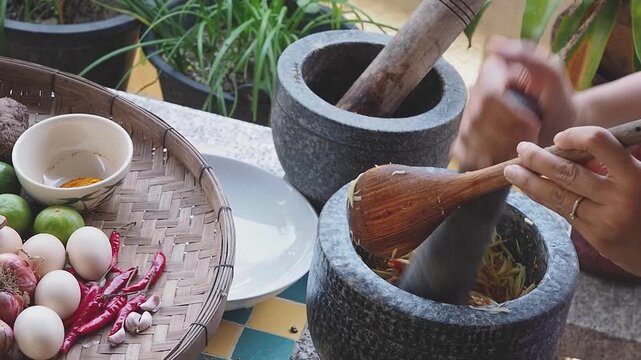 Authentic Thai cooking scene with fresh ingredients and traditional stone mortar and pestle. Hands preparing spicy sauce using chilies, garlic, lime, eggs, and herbs on a rustic kitchen table. Concept