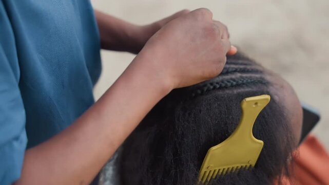 Closeup black hands parting natural hair outdoors, stylist using yellow pick comb to section scalp, patient cornrow braid being started, textured hair detail, warm sunlight, communal street salon