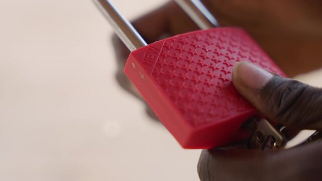 Closeup black hands holding red padlock, careful locksmith inspection of keyhole and shackle, textured plastic body, metal key turning slowly, warm natural light, shallow depth, focused deliberate
