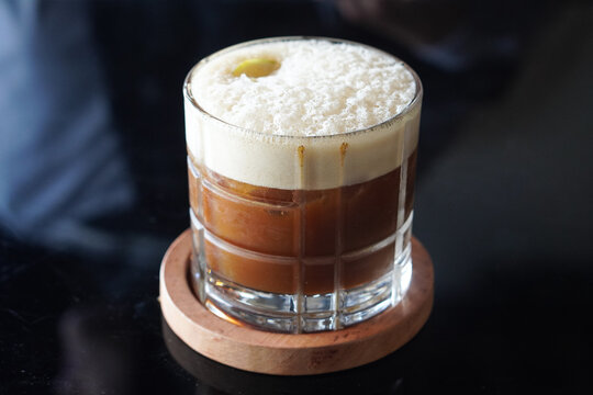 Creamy signature coffee in a clear glass with thick foam and rich brown tones in a cozy caf&eacute; setting.