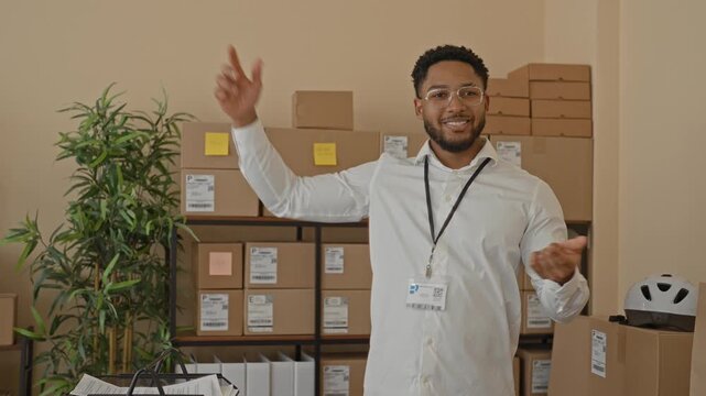 Man with parcel and visible hand framing gesture among stacked delivery boxes in a building, wearing badge and smiling; cheerful efficiency.