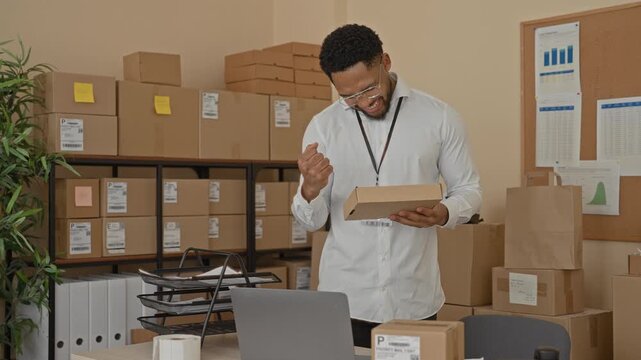 Man holding a parcel with one hand, smiling and reaching for a laptop amid stacked boxes and packing supplies in a building; satisfaction.