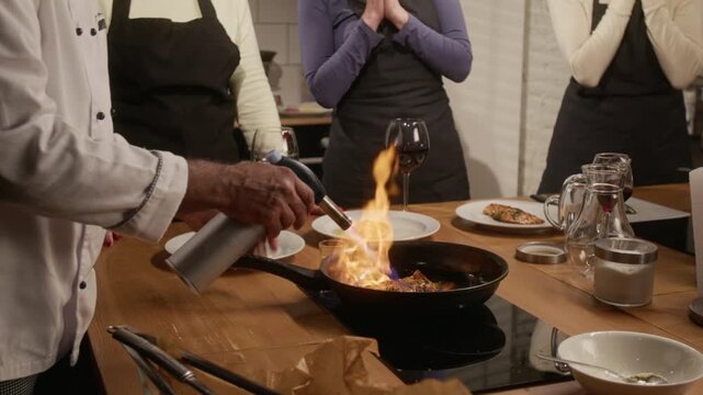 Low angle close up tilt up shot of professional chef flambeing steak for group of women at cooking master class in restaurant