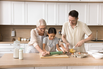 Loving multigenerational family cooking homemade pastries in kitchen, prepare dough for pizza or holiday cake. Older 60s woman enjoy time with granddaughter and grown-up son, share skills and recipe