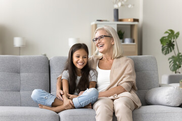 Cute school-age girl spend time with older granny, multigenerational family siting on couch, laughing, lead pleasant, trustworthy conversation, share news, joking, enjoy carefree rest at home together
