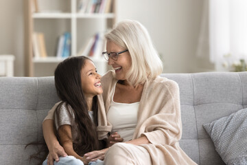 Loving attractive senior woman cuddling her adorable smiling granddaughter, multigenerational family resting on sofa, enjoy conversation, harmonic relationships and understanding on leisure at home