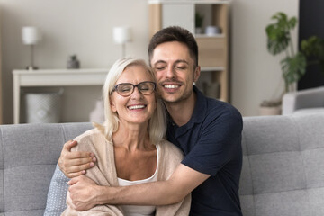 Loving young adult man embracing his older smiling mother, sit together on couch in living room, enjoy their harmonic, warm relationships and communication at grown up son visit at mommy house. Ties