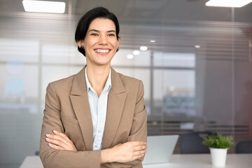 Professional office portrait of smiling Caucasian businesswoman stands with arms-crossed at corporate workplace, showing confidence, motivation and success in business and entrepreneurship, copy space