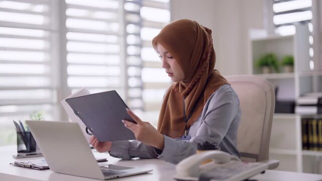 Asian Muslim professional woman reviewing business reports with laptop in modern office environment diligent focused research corporate paperwork