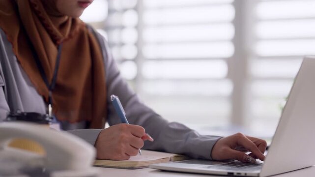 Asian adult woman professional with hijab writing notes on notebook typing laptop computer working office desk focused diligent worker productivity
