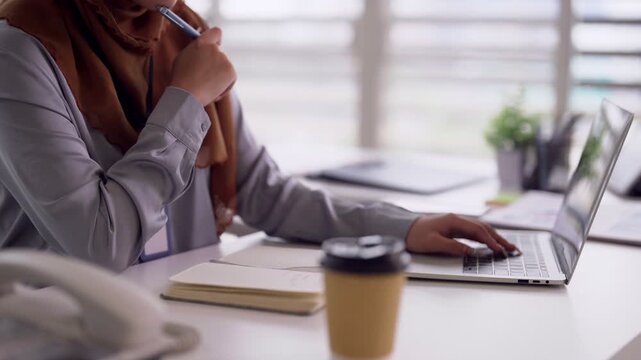 Asian adult Muslim woman professional employee writing notes creating reports typing laptop computer focused on work in modern office workspace