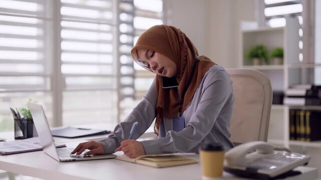 Professional Asian adult woman remote working multitasking on laptop smartphone making notes contemporary office environment