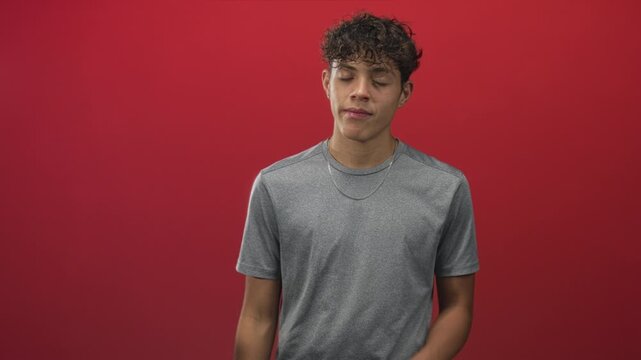 Teenage man places hand on chest with eyes closed and calm expression against red studio backdrop; sincerity gratitude.