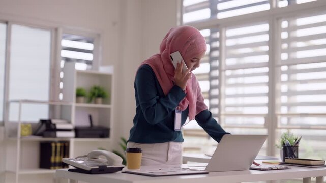 Asian Muslim adult businesswoman multitasking calling smartphone typing laptop modern home office remote work online communication