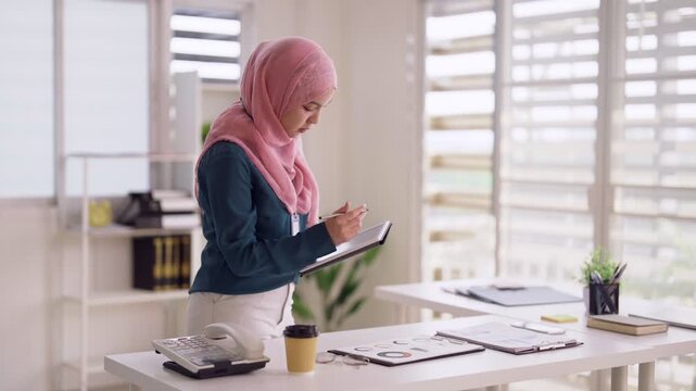 Adult Asian Muslim woman standing at office desk writing notes in notebook planning business strategy modern professional workplace environment