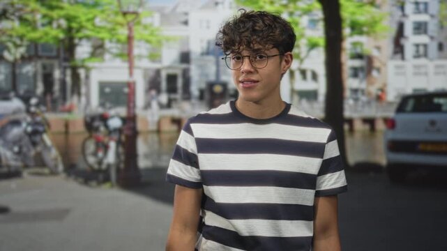 Man in striped shirt raises right hand showing bare forearm and tilts his head while standing on a city street by parked bikes and a car; contemplation.