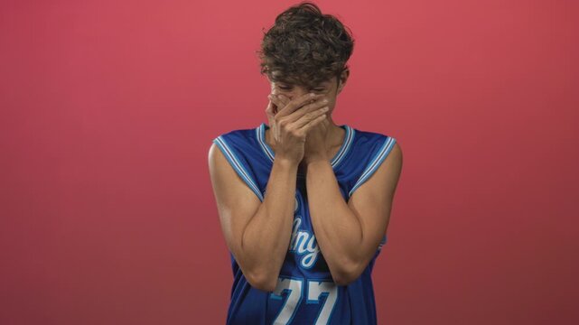 Man covers mouth with hands in studio wearing blue basketball jersey with number 77, shy grin visible; shyness amusement.