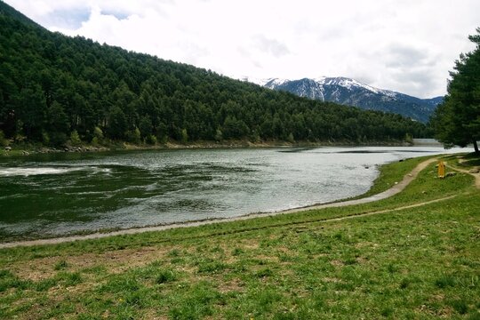 Scenic view of Lake Engolasters in Andorra featuring a mountain lake, pine forest on a slope, and snow-capped peaks under a cloudy sky. A narrow path runs along the grassy shore in the foreground area