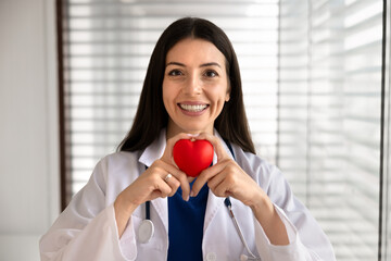 Headshot portrait smiling female doctor looking at camera, holding red heart as symbol of cardiac health awareness, represents professional cardiology services, patient care, medical checkup in clinic