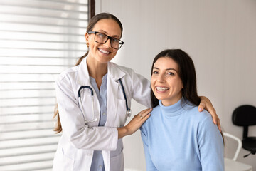 Portrait of smiling female doctor hugging on shoulder positive adult woman clinic patient during medical consultation in hospital office. Healthcare services, regular health checkup, insurance, trust