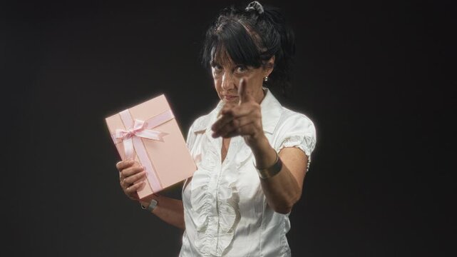 Senior hispanic woman in white blouse holding pink gift box with ribbon and points finger toward camera in studio; command attention.