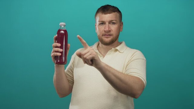 Man holding a bottle, pointing finger at the bottle and touching his chin in studio with teal backdrop; curiosity.