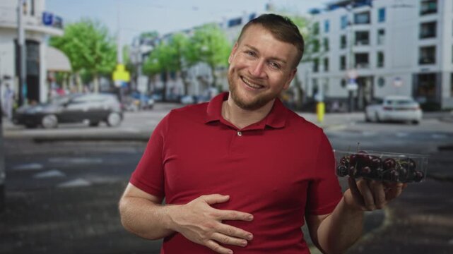 Man holding container of cherries with hand on stomach while smiling on a city street; hunger relief happiness.