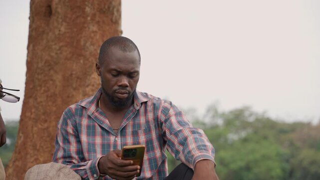 Black father and son silent pause as father checks phone, closeup on worried father scrolling messages while son watches from background mood of uncertainty, solitude under tree, technology bringing