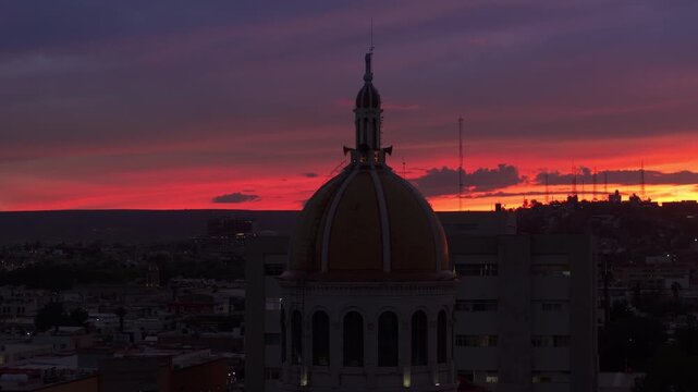 Stunning sunset timelapse over historic cathedral dome in Durango, Mexico. Vibrant orange and purple clouds paint the sky as city lights begin to glow across the urban landscape at dusk.