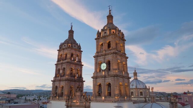 Catedral Basilica Menor de la Inmaculada Concepcion in Durango, Mexico glows warmly during sunset. Twin baroque towers with clock face rise against pink and blue evening sky with wispy clouds.