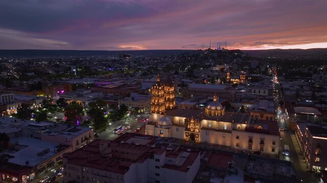 Stunning aerial view of Durango Mexico featuring illuminated cathedral with baroque towers and domes against dramatic purple and orange sunset sky as city lights twinkle across the urban landscape.
