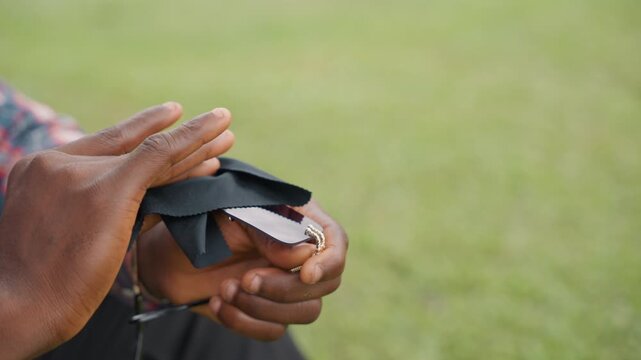 Closeup black father handing sunglasses to son on grassy bench, folded case and chain tucked inside, small gift exchange layered with memory and warmth, gentle offer and grateful receive, quiet family