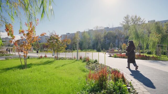 Elegantly dressed woman walks slowly along winding path in city park. Stylish female enjoys beautiful view of distant urban buildings