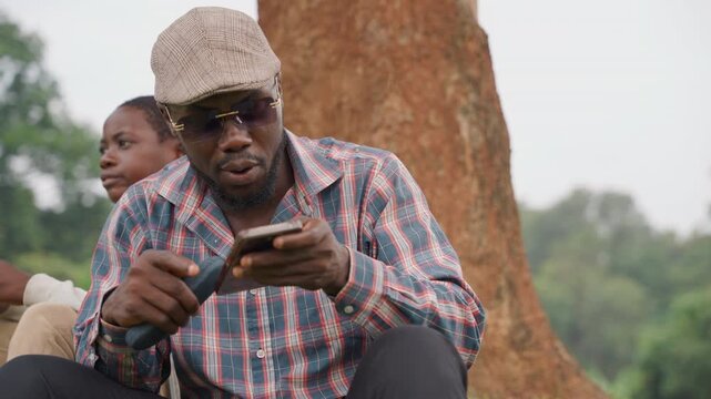 Black father and son sitting under tree examining portable speaker and phone, quiet rural park setting, father in cap and patterned shirt studies device while son watches with curiosity, warm bonding