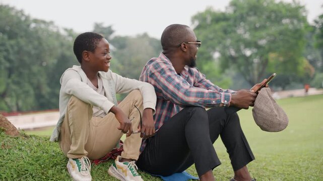 Father Son Bonding. Father and Son Relax in Green Outdoors. Casual Moment with Father and Son Sharing Laughter. Father and Son Spend Quality Time Together in Tranquil Natural Setting