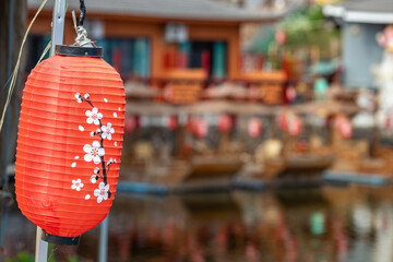 Vibrant red Japanese lantern adorned with delicate cherry blossoms, hanging against a softly blurred background of traditional buildings and their reflections on water, evoking serene cultural charm. © BestTravelPhotos
