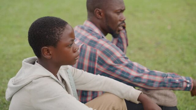 Black father and son sitting sidebyside on grass, contemplative pause as father offers silent support while son looks ahead, hooded sweater and plaid shirt, soft overcast light, peaceful park