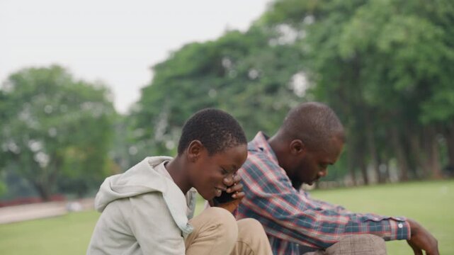 Black father and son sitting together on grass, son adjusts earbuds and laughs while father sits thoughtful, casual plaid shirt and hoodie, candid park scene showing contrast of joy and quiet support