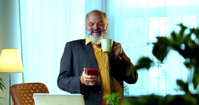 Indian senior businessman or corporate executive multitasking by speaking on a smartphone while holding a coffee or tea mug at his desk in a cozy office with professional moody lighting