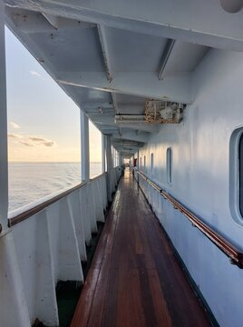 Exterior corridor of a ship deck with windows on one side and ocean view on the other side. Bright sunny day with clear sky and calm sea, travel and maritime transportation concept.