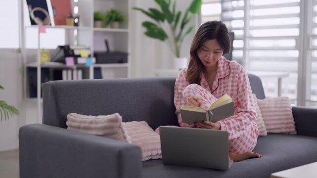 Young Asian woman remote working from home writing notes using laptop on sofa creating content enjoying comfortable lifestyle