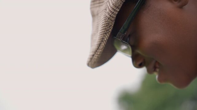 Black son closeup wearing cap and glasses, head tilted down in soft focus warm skin texture and bokeh greenery background expression of curiosity and concentration, intimate candid moment suited