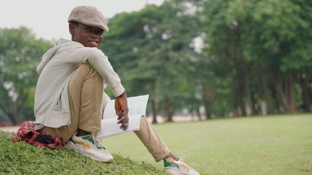 Black teenage son smiling outdoors laptop sitting on grassy hill with cap and hoodie looking at camera confident gaze creative energy casual sneakers bright daytime friendly mood ambition