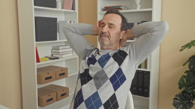 Man in argyle sweater leans back in chair with hands behind head in an office building near bookshelf and plant; relaxation reflection.