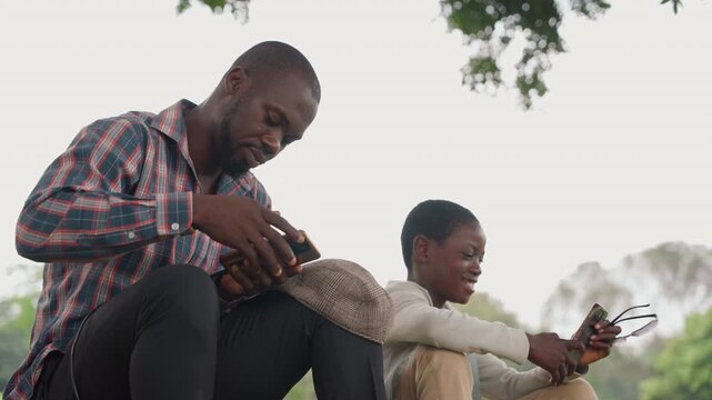 Black father and son using device, father demonstrates controller while son watches eagerly, outdoor lesson under tree, candid smiles, drone or smartphone interaction, playful learning, modern