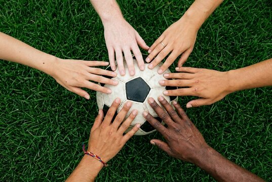 Six diverse hands of different skin tones touching soccer ball on green grass symbolizing unity and global connection