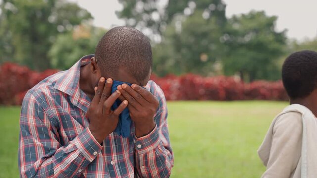Black father wipes face with cloth in park while son stands nearby, emotional pause as handkerchief meets wet cheeks, plaid shirt and greenery backdrop, father composing after tearful moment, quiet
