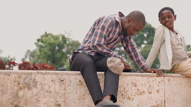 Black father and son sitting on stone ledge in park, father in plaid shirt gently pats sons head while reaching into woven bag warm daylight, calm conversation, intimate caring exchange, mentorship