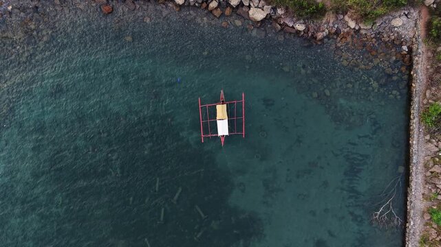 Top down ascending drone shot above a traditional Filipino bangka outrigger boat anchored in clear shallow water along a rocky shoreline near Puerto Princesa Palawan Philippines