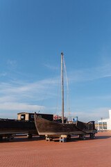 Fototapeta premium Old wooden sailing boat rests on supports outside on a paved pier area under a clear blue summer sky waiting for restoration work to begin in the coming days